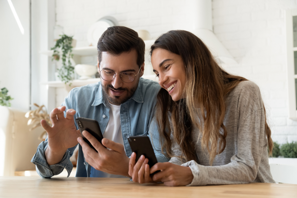 Young happy couple using two phones share social media news at home